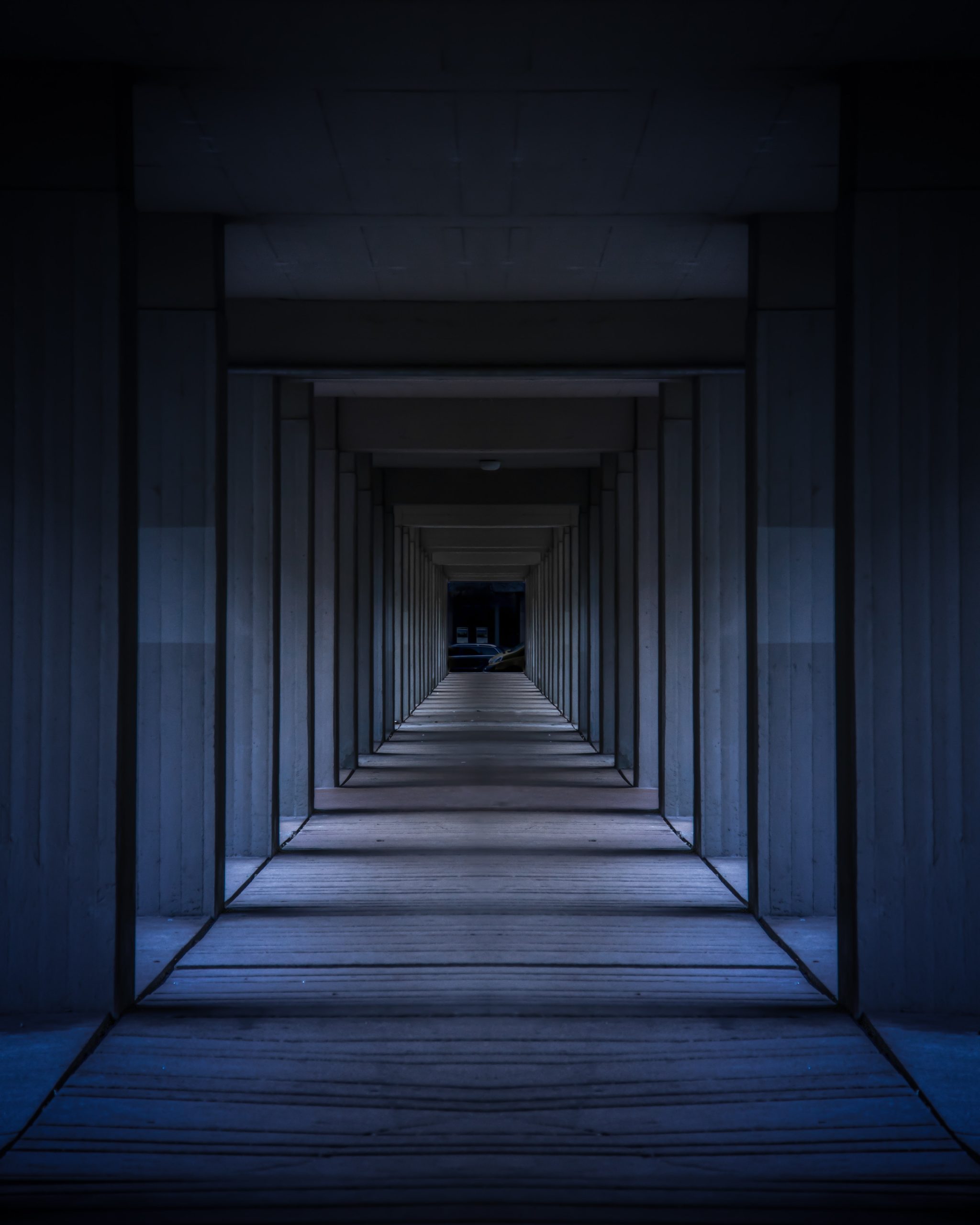 Long couloir à colonnes de béton dans la pénombre bleue, perspective symétrique vers une ouverture sombre, image d'une architecture invisible.
